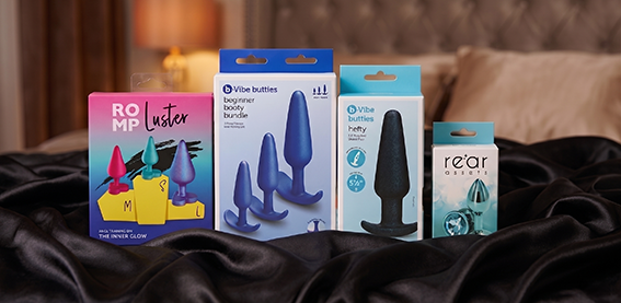 Neljä erilaista anaalitappi-tuotepakkausta aseteltuna vierekkäin mustalle, silkkiselle kankaalle sängyn päälle. Vasemmalta oikealle: värikäs ROMP Luster -pakkaus, sininen b-Vibe booties Beginner booty bundle -setti, vaaleansininen b-Vibe booties Hefty -pakkaus ja pienempi Rear Assets -koristetulpan pakkaus. Taustalla näkyy pehmeästi tarkennettu makuuhuoneen sisustus ja sängynpääty.